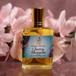 A small rectangular glass perfume bottle filled with golden liquid stands in front of soft pink flowers, with a bright blue label that reads “Cherry Blossom Sacred Perfume” and a shiny gold cap on top.