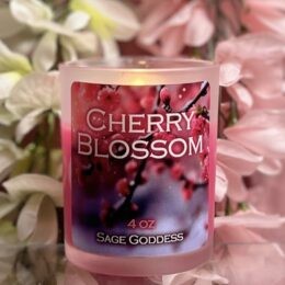A lit pink glass candle labeled “Cherry Blossom” and “Sage Goddess 4 oz” sits in front of a close up image of cherry blossoms, surrounded by soft pink and white flower petals in the background.