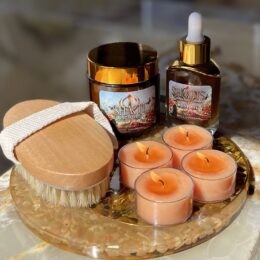 Four lit orange tealights, one wooden body brush, one dropper bottle, one amber cosmetic jar on stone plate.