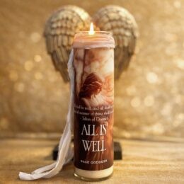 A tall glass jar candle with a soft flame at the top sits in front of gold metallic angel wings. The label shows a sepia image of a robed figure and the words “ALL IS WELL” above the Sage Goddess logo. A white fabric ribbon hangs from the neck of the candle, and the background is an out of focus gold surface.