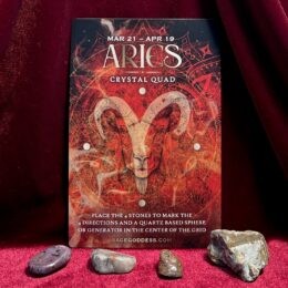 A red Aries themed crystal grid card with a white ram’s head at the center, surrounded by fiery geometric patterns, with four small stones placed in front on red fabric.