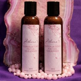 Two pink-labeled bottles of “Athena’s Exfoliating Facial Cleanser” stand upright on a polished pink agate slice, surrounded by pale pink beads against a purple cloth background.