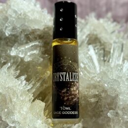 A 10 ml Crystalize perfume oil by Sage Goddess in a glass roller bottle with a black cap, standing among clear quartz crystal points on a soft, neutral background.