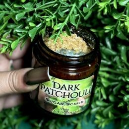 Dark Patchouli Scrub jar open with golden scrub visible and tumbled labradorite visible. Foliage background.