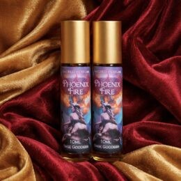 Two gold-capped roller bottles of “Phoenix Fire” sacred perfume stand side by side against rich gold and crimson velvet fabric, with a fiery phoenix-themed label in vivid colors.