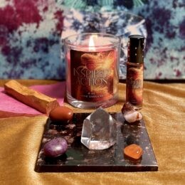 A lit glass candle labeled “Inspired Action” sits at the center of the image, with a matching perfume roller bottle standing beside it on the right. In front of them is a small rectangular stone slab holding a clear quartz point and several colorful tumbled stones in orange, red, purple, and white. A piece of palo santo wood lies to the left on pink tissue, and the whole scene rests on a golden cloth with a soft, tie-dyed, blue-and-purple fabric backdrop.