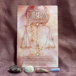 A pink-toned “Full Moon in Libra Crystal Quad” card with a golden Libra scales design stands upright against a mauve fabric backdrop. In front of the card, four small polished stones in shades of dark plum, soft white, dusty pink, and light blue are lined up in a row.
