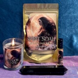 Photo of a lit “Shadow Rising” candle, a “Spirit Soak” bath crafting set, a perfume roller, and a long black double-terminated crystal displayed together on colorful fabric.
