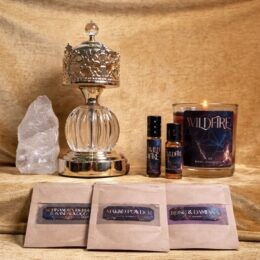 A gold velvet backdrop displays a small altar-like arrangement. On the left is a tall, rough clear quartz point. In the center stands an ornate, crown-topped gold and glass incense burner. To the right are two small Wildfire-branded perfume bottles in dark glass, and a lit Wildfire candle in a clear glass jar with a dark cosmic-style label. In front, three flat brown paper packets with matching dark labels are laid in a row, containing different ritual powders or blends.