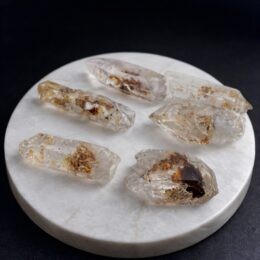 Six clear quartz points with golden mineral inclusions rest on a round white stone slab against a dark background.