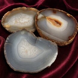 Three polished gray and white agate slices with natural banding and crystalline centers rest on a deep burgundy velvet fabric, showing their organic shapes and textured stone edges.