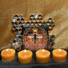 Product photo showing four lit honey yellow tea light candles in a row on black fabric, with a Queen Bee Tealights label featuring a crowned bee and laurel wreath, set against a warm golden backdrop with a metallic honeycomb decoration and a small bee figurine beside the label.