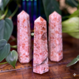 Three polished pink-orange crystal generators with pointed tips stand upright on a wooden surface, their sparkling mottled texture framed by soft green leaves in the background.
