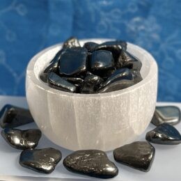 White selenite bowl filled with shiny black tumbled stones, with more matching stones scattered around the base, set against a soft patterned blue background.
