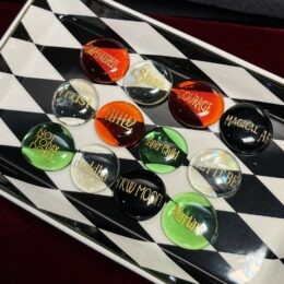 A rectangular dish with a black and white diamond pattern holds a grid of round glass stones in red, green, black, and clear colors, each etched with gold words like Empowered, Shine, Courage, Magical AF, Trust, Wild, Inner Child, Not Today Doris, Buddha, New Moon, Let It Be, and Nurture.