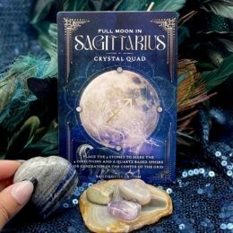 Full Moon in Sagittarius Crystal Quad card displayed behind four tumbled gemstones arranged on an agate slice, held against a teal feather and sequin background.