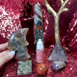 Hand holding a carved crystal moon among Root Chakra tuning drops, crystal tower, pyramid, and stones arranged on a red sparkling altar cloth.