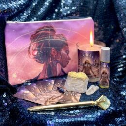 A Gemini New Moon altar set features a cosmic goddess pouch, a lit Sacred Song candle, a roller perfume, a gold pen topped with green fluorite, mantra cards, and sodalite, blue lace agate, and clear quartz stones arranged on vibrant blue fabric.