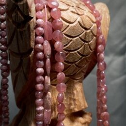 Strands of polished pink rhodochrosite beads in various shapes drape over a carved wooden owl, highlighting their glossy rose color against the textured wood.