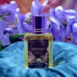 A square glass bottle of Sage Goddess “Downshift” nervous system perfume with a purple cap sits on teal fabric in front of soft purple flowers.