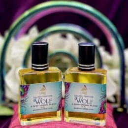 Two bottles of Sage Goddess “Wolf” sacred perfume stand on a dark purple surface in front of an iridescent rainbow arch and soft white flowers.