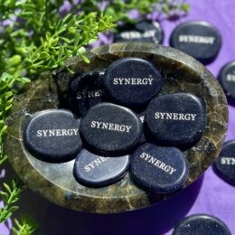 A small polished blue goldstone palm stone, engraved with the word “SYNERGY,” rests in a bowl among similar stones, sparkling under bright light.