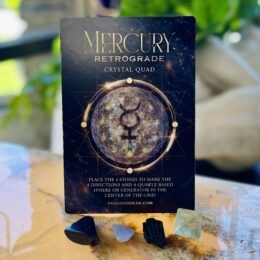 A Mercury Retrograde Crystal Quad card with a cosmic Mercury symbol is displayed behind four small stones arranged on a golden, marbled surface.