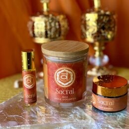 An orange Sacral Chakra collection featuring a candle, perfume oil, and balm is arranged on a reflective surface with golden decor behind.