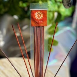 A clear package of orange Sacral incense sticks stands upright, surrounded by several loose incense sticks fanned out in front.