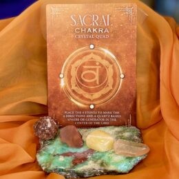 An orange Sacral Chakra Crystal Quad card stands behind four small tumbled stones arranged on a green crystal slab.